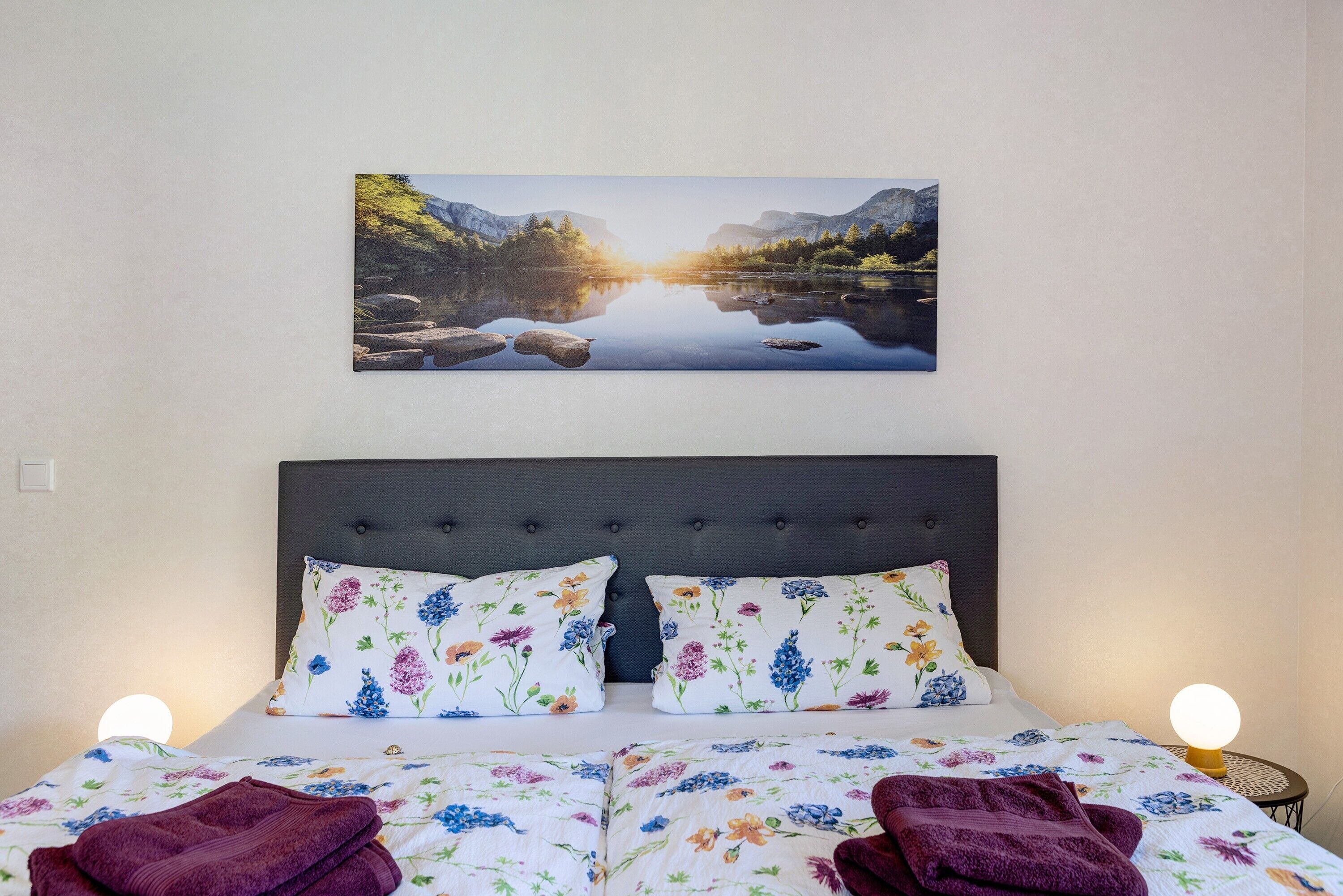 Un lit avec une literie à motifs floraux, une tête de lit et un tableau représentant un lac accroché au mur.