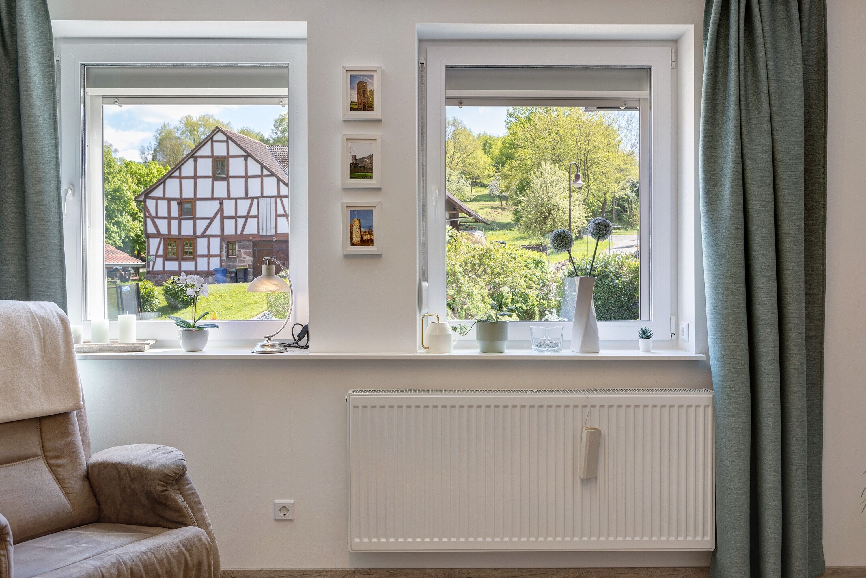 Ein Zimmer mit zwei Fenstern, einem Heizkörper, einem Stuhl und einem Regal mit Topfpflanzen und gerahmten Bildern.