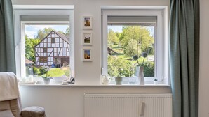 Une pièce avec deux fenêtres, un radiateur, une chaise et une étagère où sont posées des plantes en pot et des photos encadrées.