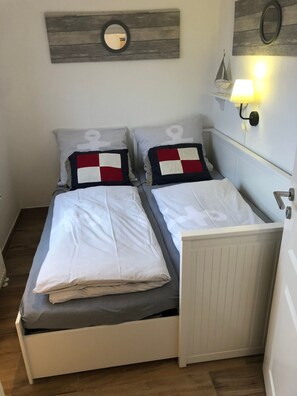 A double bed with a grey and white striped duvet, two pillows with a red and white pattern, a small round mirror, and a wall-mounted lamp.