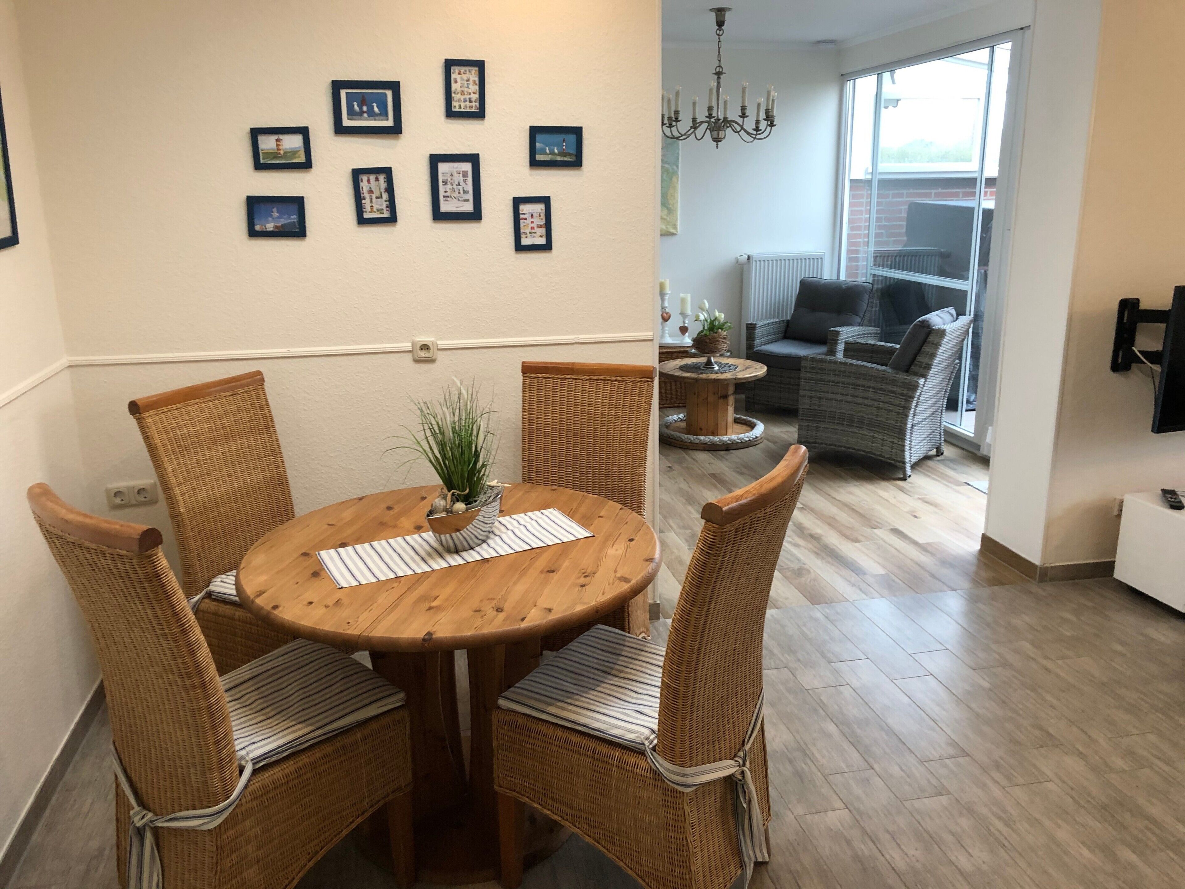 A dining area with a round wooden table, four wicker chairs, a potted plant, and framed pictures on the wall.