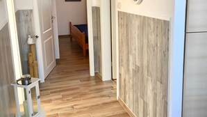 A hallway with wooden flooring, a bed in the background, a white door, and a small table with a pot on it. - Holiday Home 'In Strandnähe' with Private Terrace, Private Garden and Wi-Fi (Dornum)