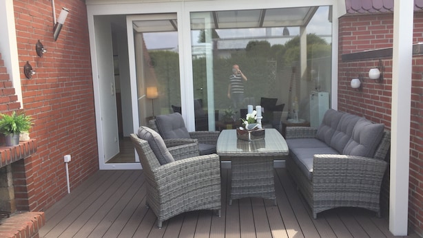 A covered patio with wicker furniture, a glass door leading to a garden, and a brick wall. - Holiday Home 'In Strandnähe' with Private Terrace, Private Garden and Wi-Fi (Dornum)