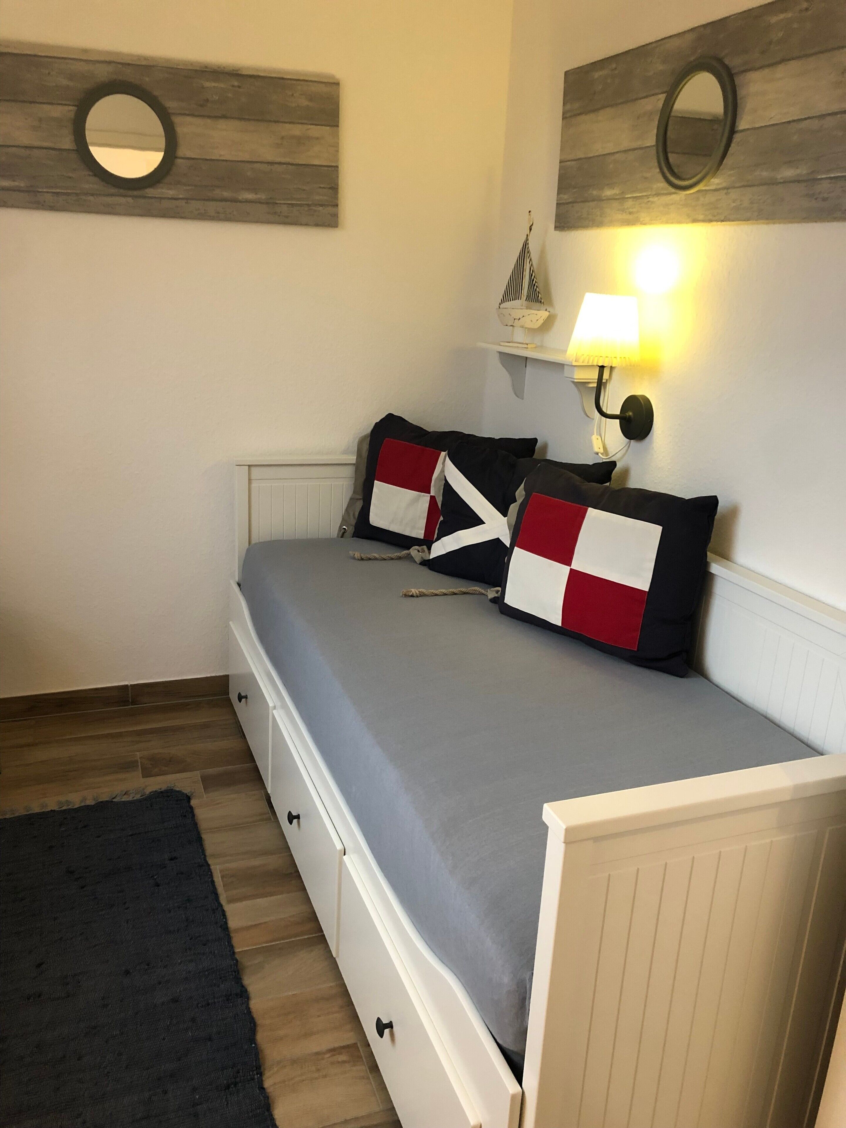 A bedroom with a bed, a nightstand, a lamp, a wall-mounted mirror, and a small shelf with a decorative item.