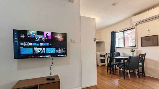 A living room with a flat-screen TV mounted on the wall displaying various streaming services. There is a wooden cabinet with a remote control on top, a dining area with a table and chairs, and a window with curtains. - 15 mins to Airport|Gateway to NSW (Campbellfield)