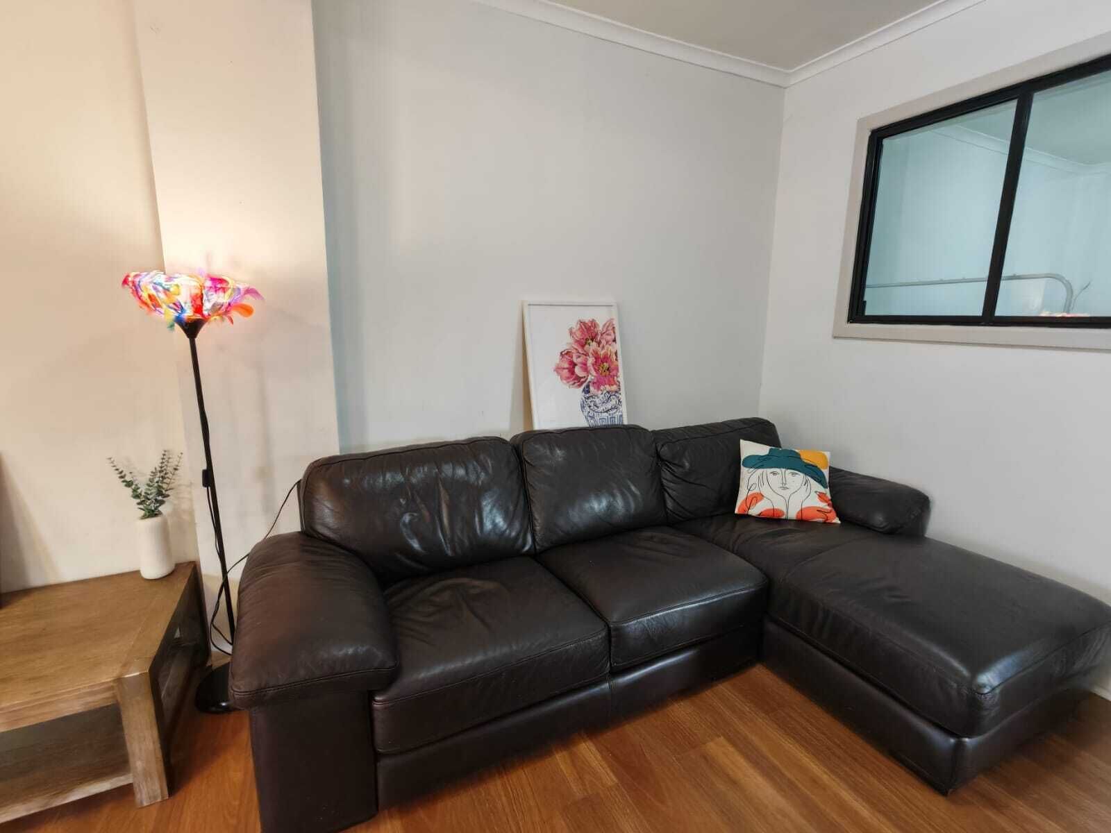 Um sofá de couro preto numa sala de estar com chão de madeira, uma luminária de pé e um quadro emoldurado na parede.