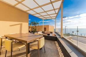 Een moderne eethoek buiten met uitzicht op zee, een glazen balustrade en een zithoek.