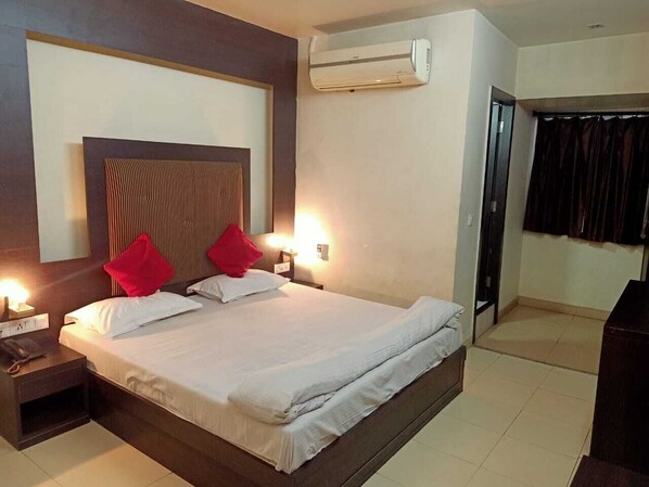 A hotel room with a large bed, a bedside table with a lamp, an air conditioner, and a view of the outside.