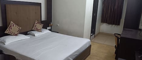 A hotel room with a bed, a desk, an air conditioning unit, a door, and a curtain.