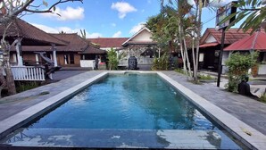 Ein rechteckiger Pool mit Fliesenrand, umgeben von einem gepflasterten Bereich mit Palmen und Gebäuden im traditionellen Stil mit roten Dächern.