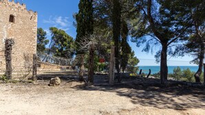 Un edificio histórico de piedra con fachada robusta, rodeado de árboles y una vista despejada al mar.