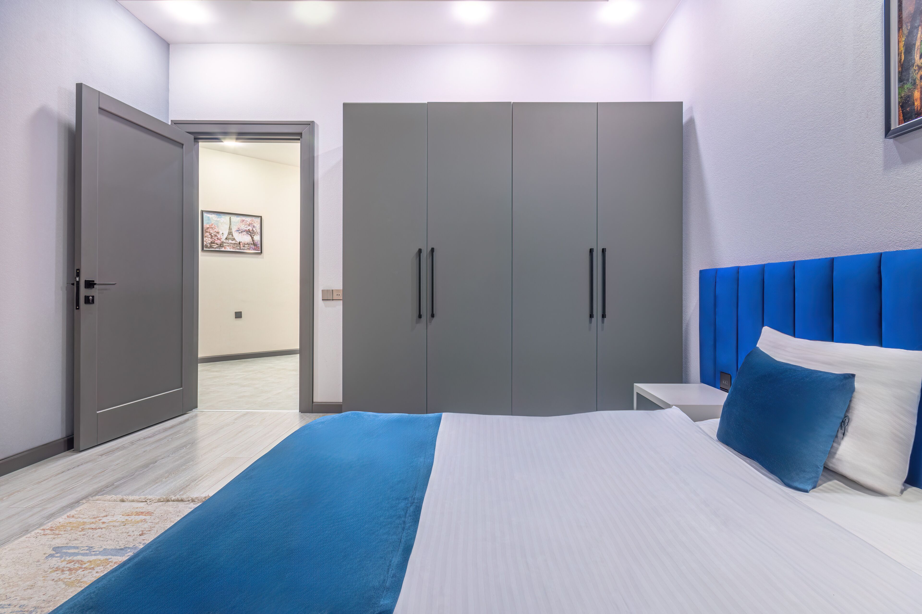 A modern bedroom with a bed, a nightstand, a wardrobe, and a door leading to another room.