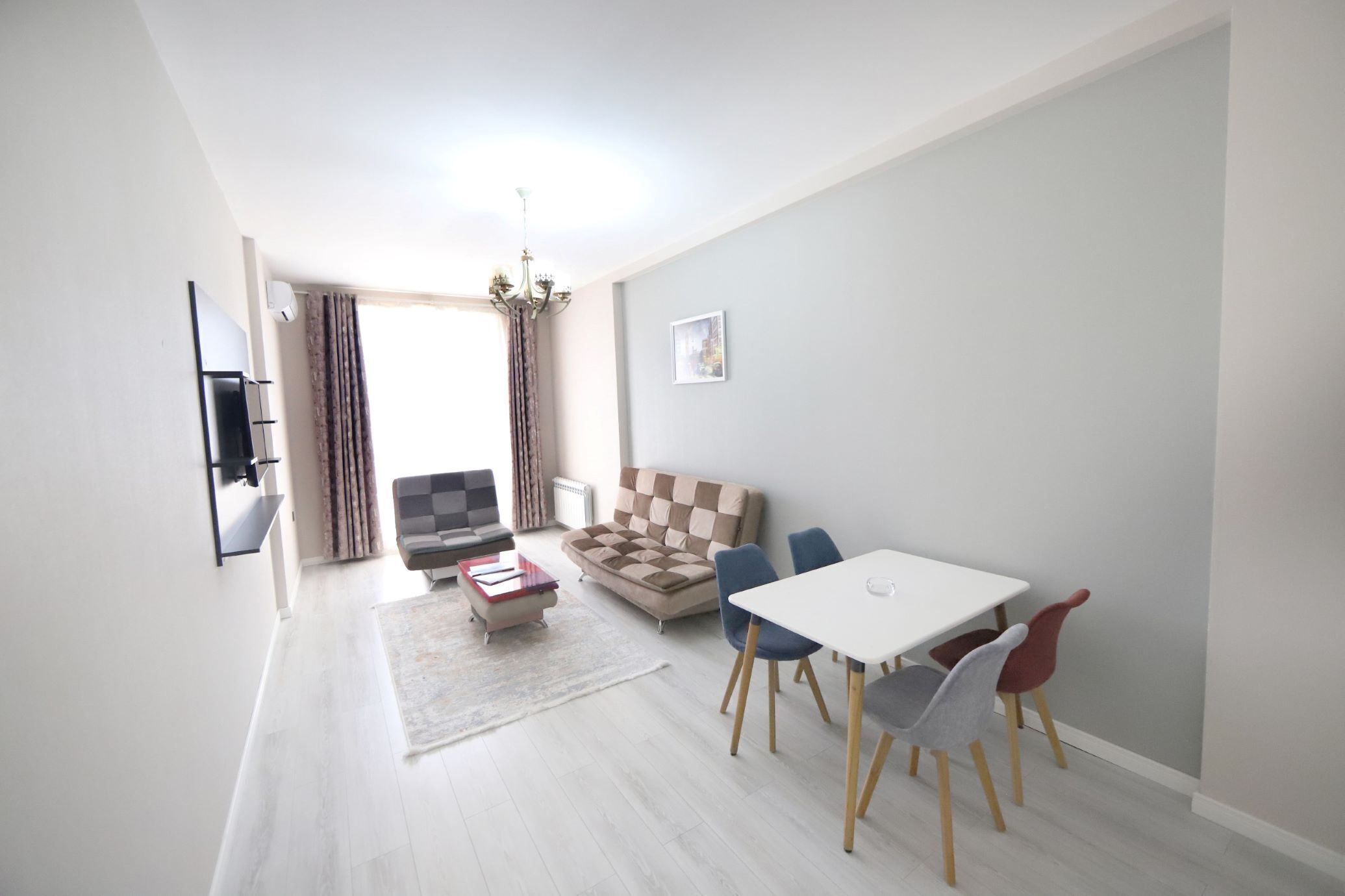 Una sala de estar moderna con sofá, una mesa de comedor con sillas, un televisor montado en la pared y una araña.