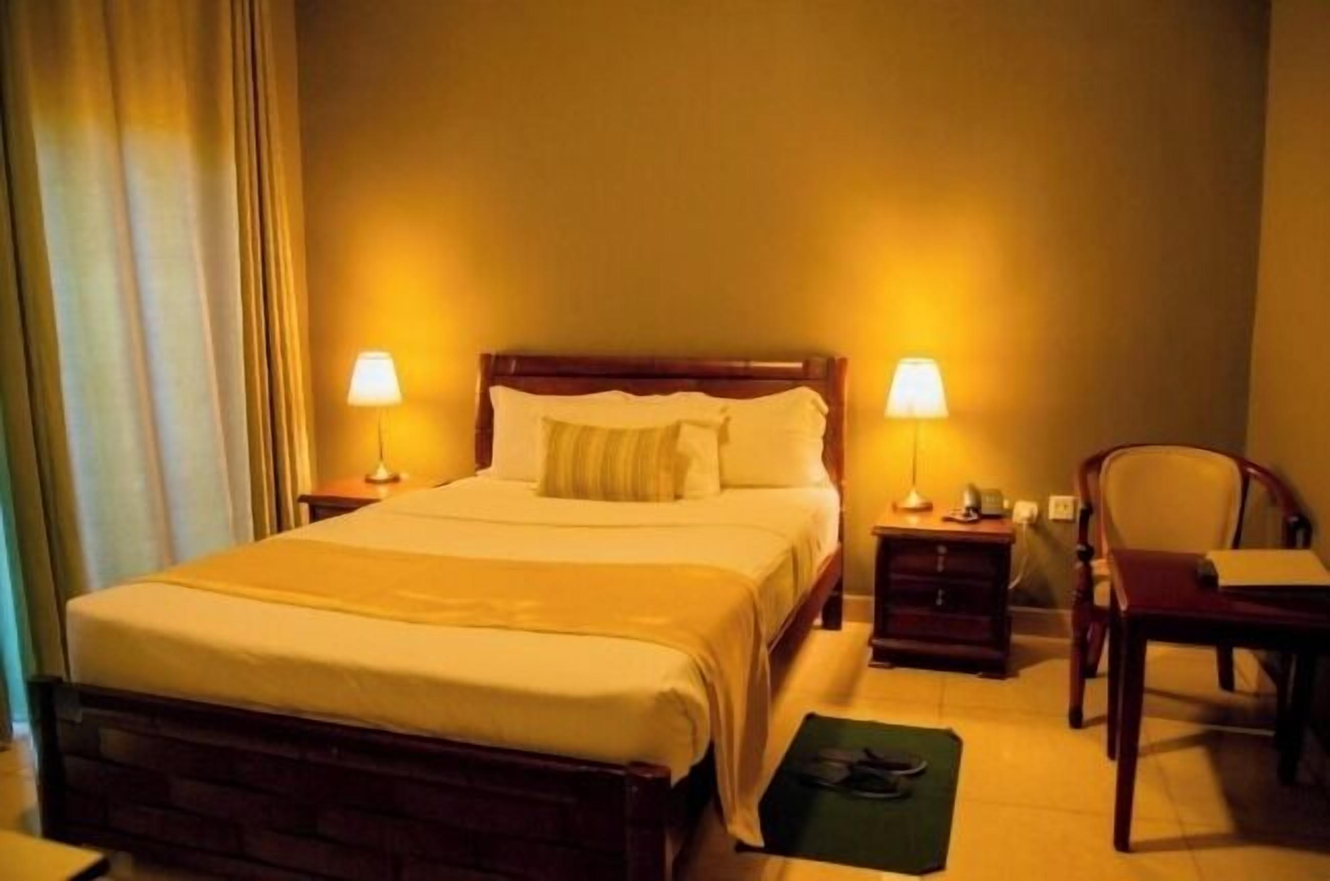 A hotel room with a large bed, two bedside lamps, a wooden nightstand, a chair, and a desk with a lamp.