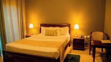 Habitación de hotel con una cama grande, dos lámparas de noche, una mesita de noche de madera, una silla y un escritorio con una lámpara.