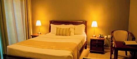 A hotel room with a large bed, two bedside lamps, a wooden nightstand, a chair, and a desk with a lamp.