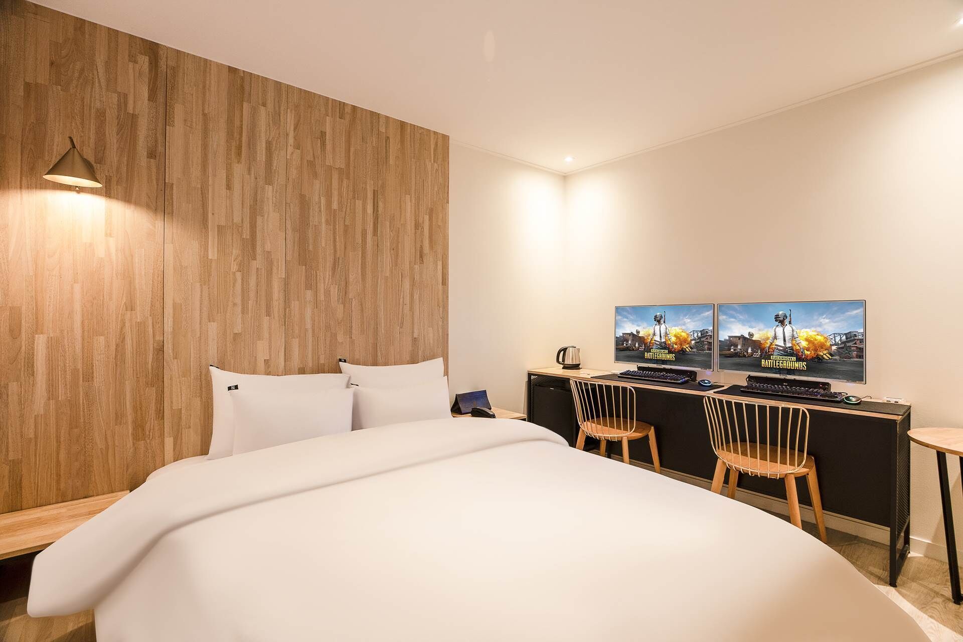 Une chambre d’hôtel moderne avec un mur d’accent en bois, un grand lit, un bureau doté de deux écrans et une chaise.
