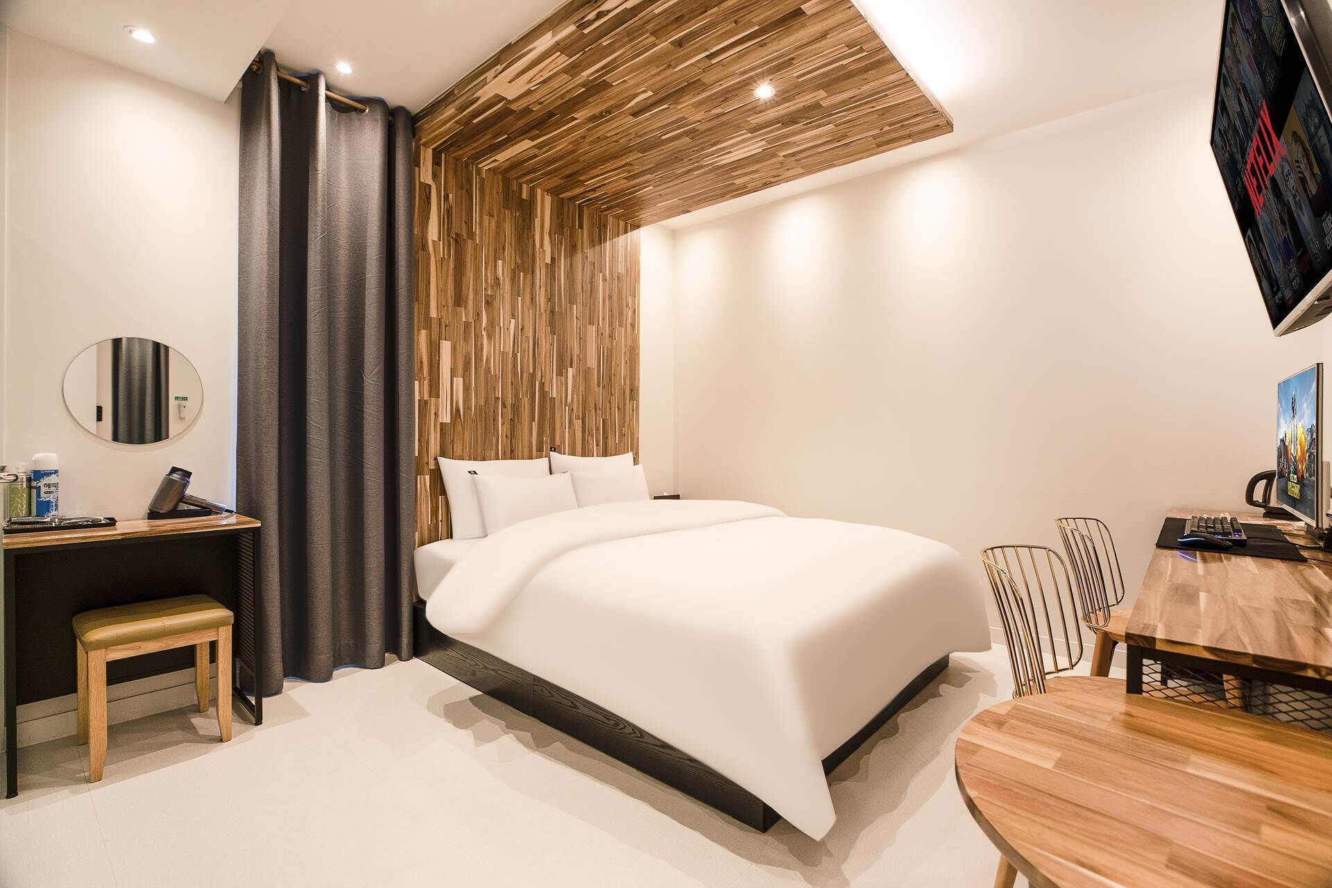 A modern hotel room with a large bed, a desk with a chair, a round table with chairs, a TV, and a wooden ceiling design.