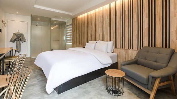 A modern hotel room with a large bed, a grey armchair, a small round table, and a wooden accent wall. - Hwagokhoteln (Seoul)