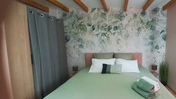 A bedroom with a bed, pillows, and a nightstand. The room has a floral wallpaper and wooden beams on the ceiling.