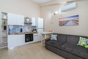 Une cuisine moderne avec des meubles blancs, un pare-feu en carrelage et un coin repas agrémenté d’un canapé gris et d’un coussin à motifs de cactus.