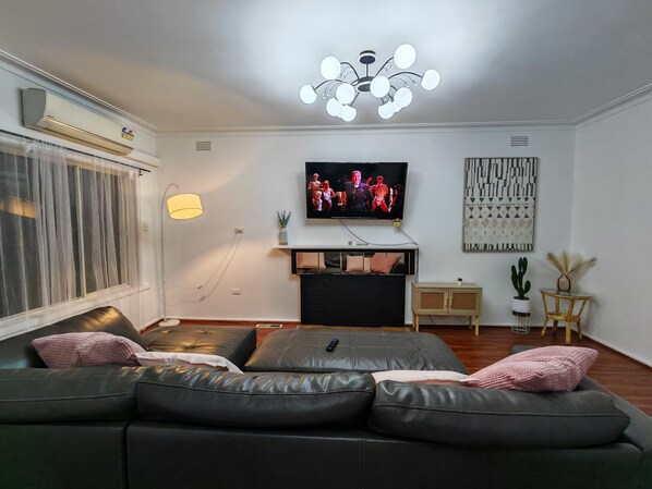 Ein modernes Wohnzimmer mit einer schwarzen Ledersofa, einem an der Wand montierten Fernseher, einer Kristallleuchtre und einem Fenster mit durchsichtigen Vorhängen.