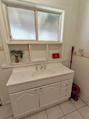 Ein Badezimmer mit einem weißen Waschbecken, einem Fenster über dem Spiegel, einer Topfpflanze, einer roten Erste-Hilfe-Ausrüstung und einem Mop auf dem Boden.