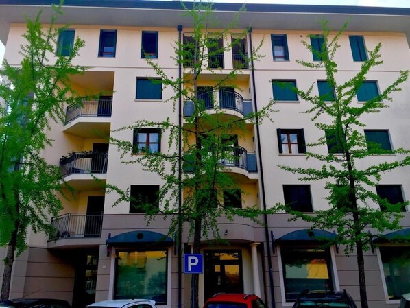 A multi-story building with balconies, a parking sign, and trees in front.