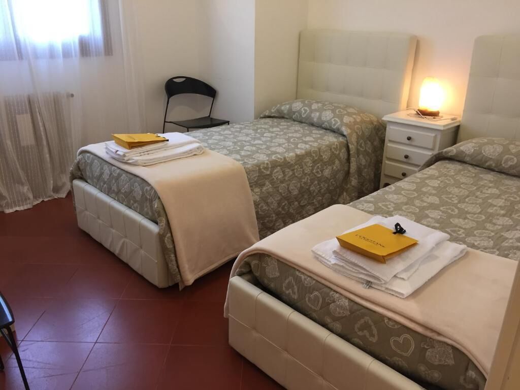 Hotelkamer met twee bedden, een stoel, een nachtkastje met lamp en een raam met dunne gordijnen.