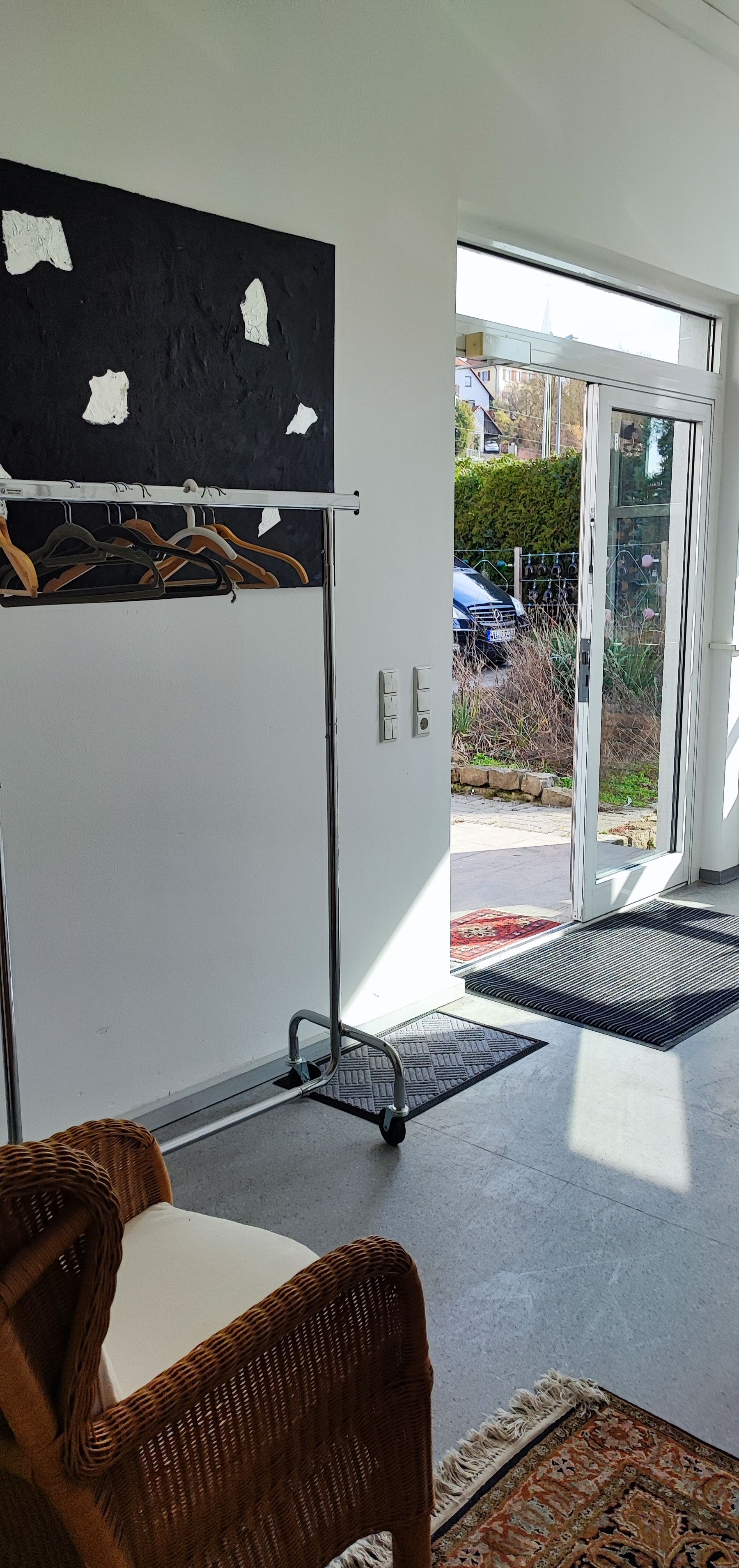 A room with a wicker chair, a coat rack with hangers, a door leading outside, and a mat at the entrance.