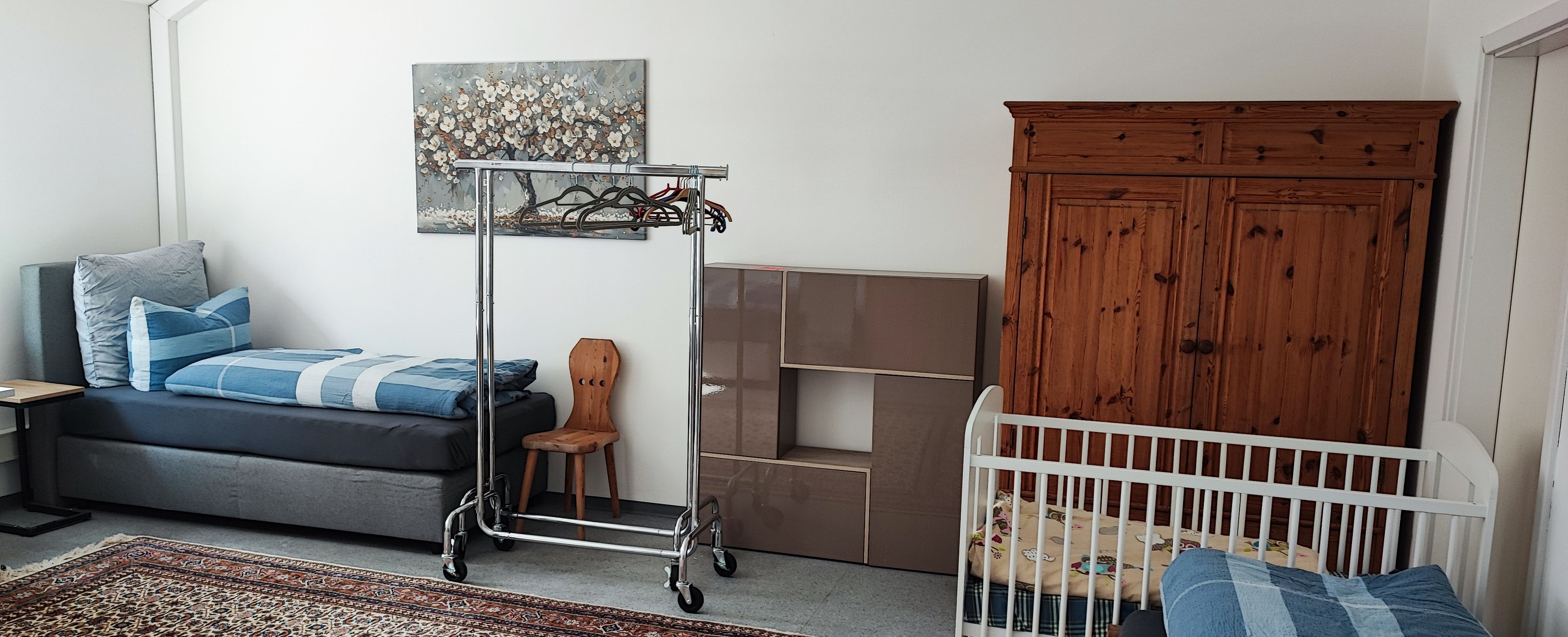 Ein modernes Wohnzimmer mit grauem Sofa, einem weißen Babybett, einem Holzschrank und einem Metallwagen.