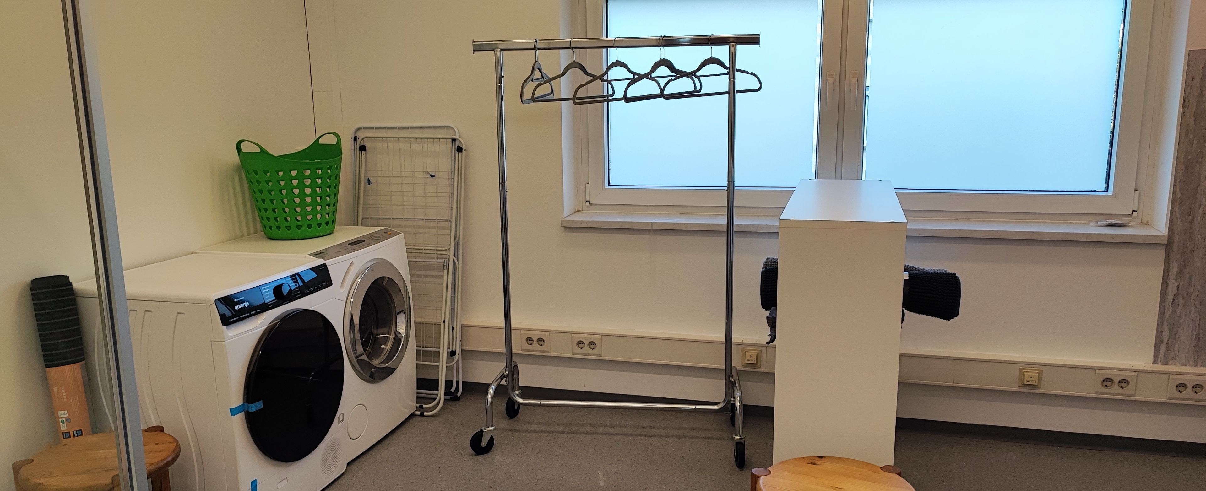 A laundry room with a front-loading washing machine, a drying rack, a green laundry basket, and a wooden stool.
