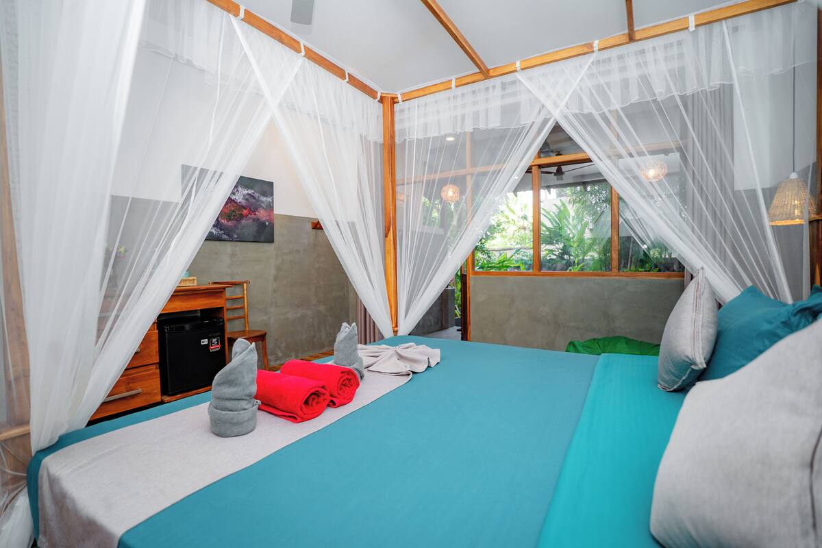 A neatly made bed with a teal bedspread and white net curtains, a small wooden table with a black appliance, and a green pillow on the bed.