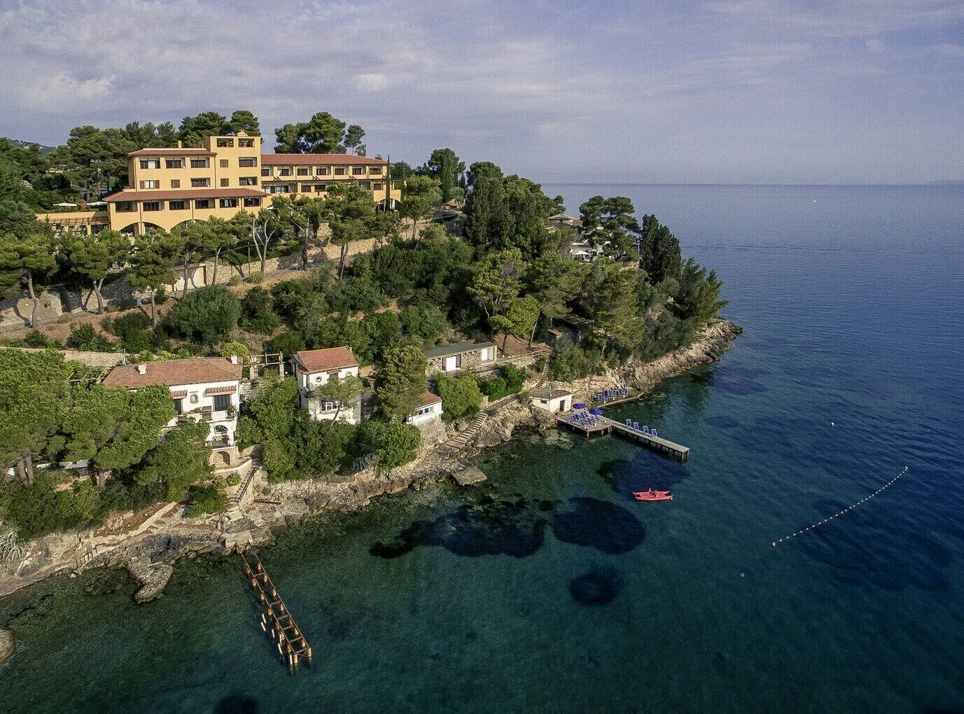 Vue aérienne d’une propriété située sur la côte, avec un grand bâtiment, des maisons plus petites et une jetée qui s’avance dans la mer.