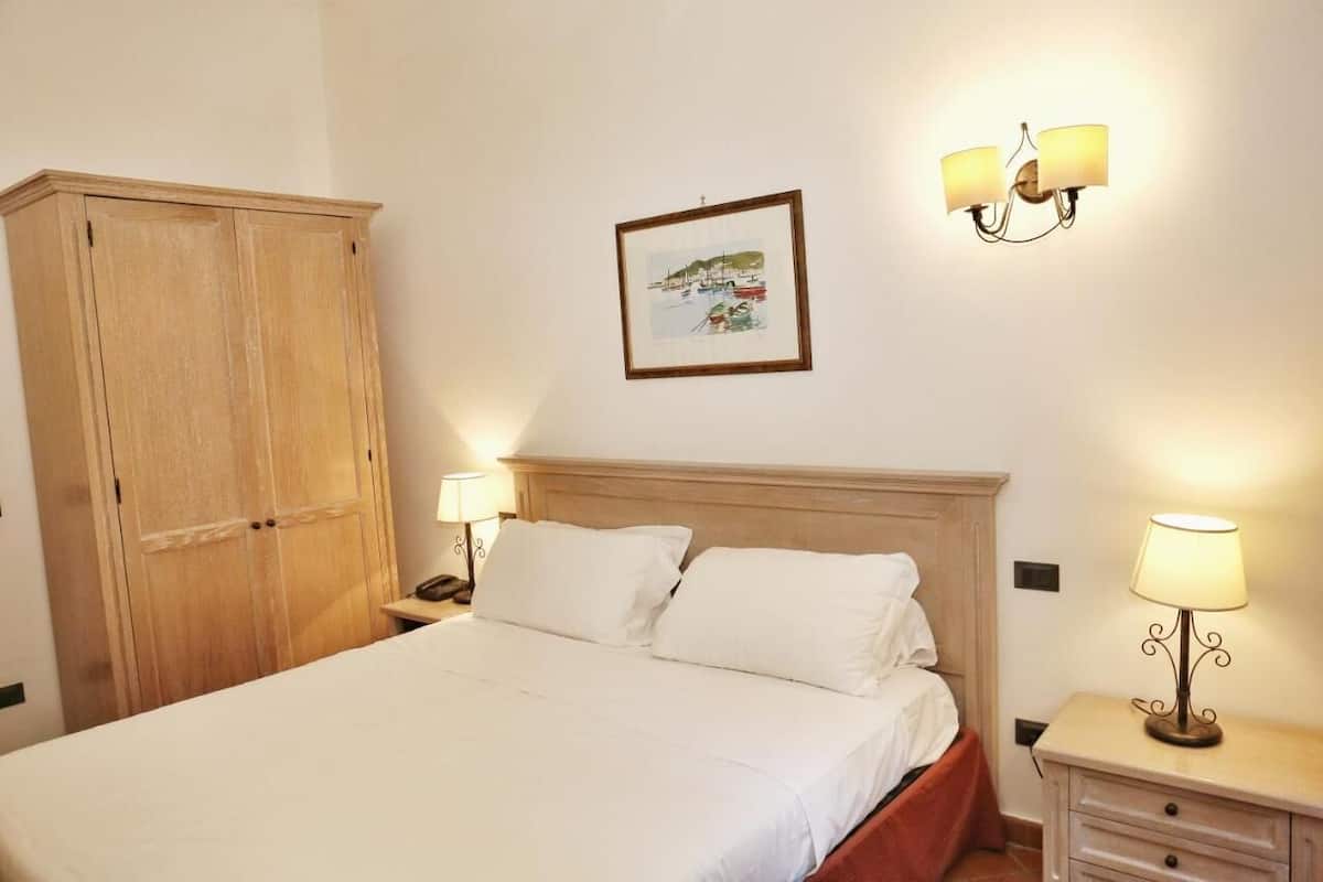 A bedroom with a large wooden wardrobe, a bed with white bedding, two bedside lamps, and a framed picture on the wall.