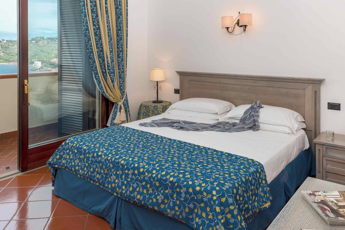 A bedroom with a bed, a nightstand, a lamp, and a view of the sea.