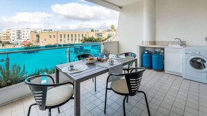 A balcony with a table and chairs, a washing machine, and a view of a swimming pool and buildings. - Seaside comfort in the heart of Alghero (Alghero)