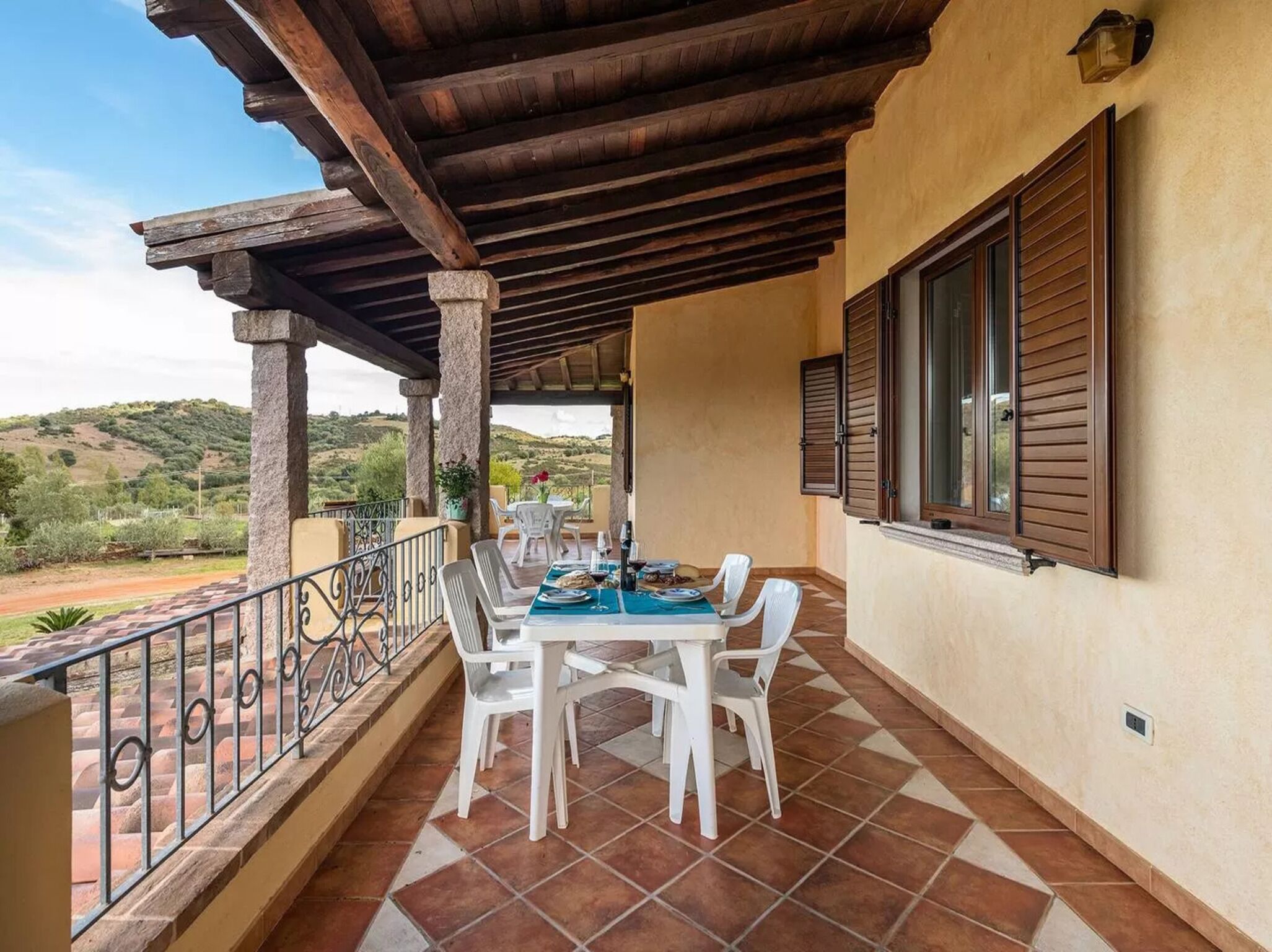 Un área de comedor al aire libre cubierta con una mesa preparada para dos personas, rodeada de sillas blancas, en una terraza con vistas a las colinas.