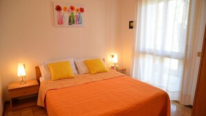 Une chambre à coucher avec une couvre-lit orange, deux coussins jaunes, un tableau de fleurs accroché au mur et une fenêtre ornée de rideaux légers.