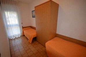 Ein Zimmer mit zwei Einzelbetten, einem Holzschrank, Fliesenboden und einem Fenster mit durchsichtigen Vorhängen.