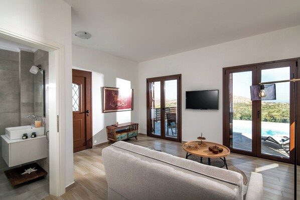 A modern living room with a sofa, a round table, and a TV. There is a sliding glass door leading to a pool area.