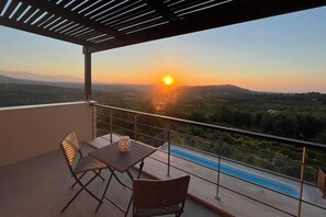 A balcony with a table and chairs, overlooking a swimming pool and a scenic landscape during sunset.