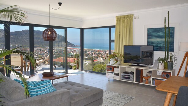 A spacious living room with a large TV, a comfortable sofa, and a view of the city and sea through large windows. - Spectacular house overlooking False Bay. (Cape Town)