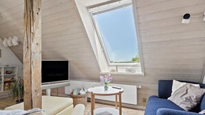 A cozy loft bedroom with a sloped ceiling, a large skylight, a flat-screen TV, a round wooden table, a blue sofa, and a bookshelf. - Cozy apartment in Nordborg (Nordborg)