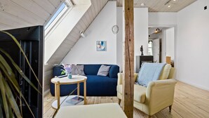 A cozy living room with a blue sofa, a small round table, and a yellow armchair. A fireplace is visible in the background, and there is a wooden ceiling with a skylight. - Cozy apartment in Nordborg (Nordborg)