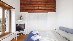 A bedroom with a wooden ceiling, a bed with white and blue bedding, a TV on a white stand, and a window with a view of greenery. - Stunning private villa for 4 guests with WIFI, TV, terrace, pets allowed and panoramic view (Locarno)