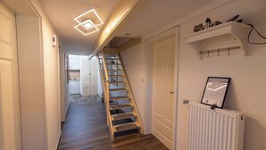 Un couloir avec un escalier en bois, un radiateur blanc et une étagère fixée au mur, sur laquelle sont posés divers objets.
