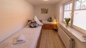 Une petite pièce bien éclairée, avec deux lits simples, un radiateur, une fenêtre avec des stores et un luminaire fixé au mur.