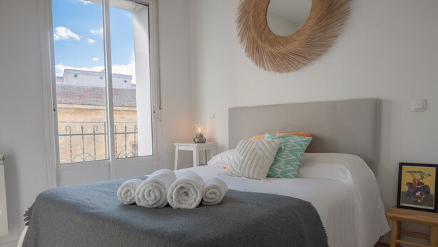 A bedroom with a bed, a bedside table with a lamp, a framed picture, and a view of the outside. - Urban Spring (Madrid)