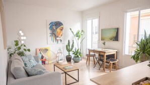 A modern living room with a sofa, coffee table, and dining area. There are potted plants and artwork on the walls. - Urban Spring (Madrid)